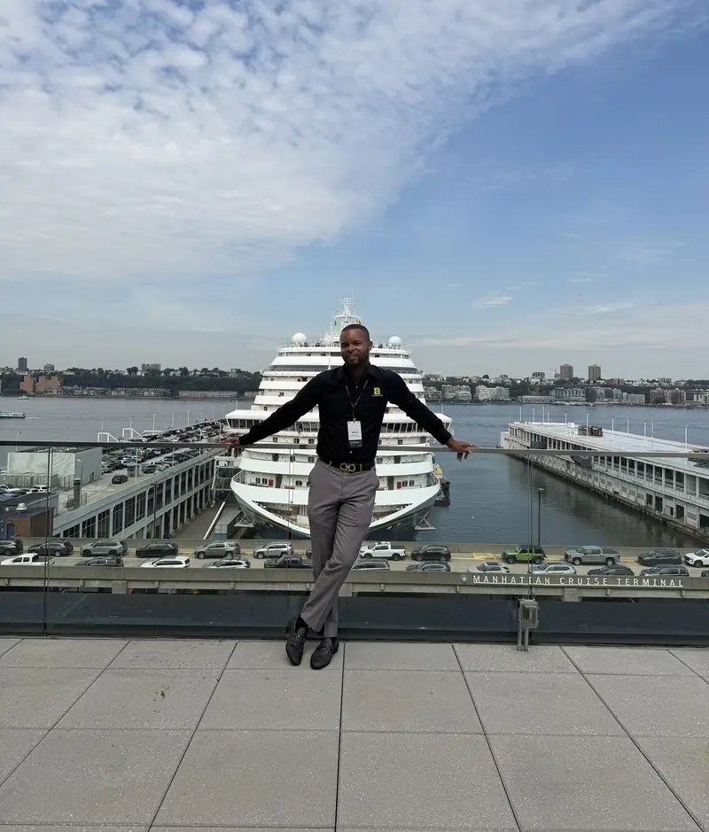 Merrick Lee Jr. sitting on a rooftop ledge with arms outstretched, a cruise ship docked behind him under a blue sky