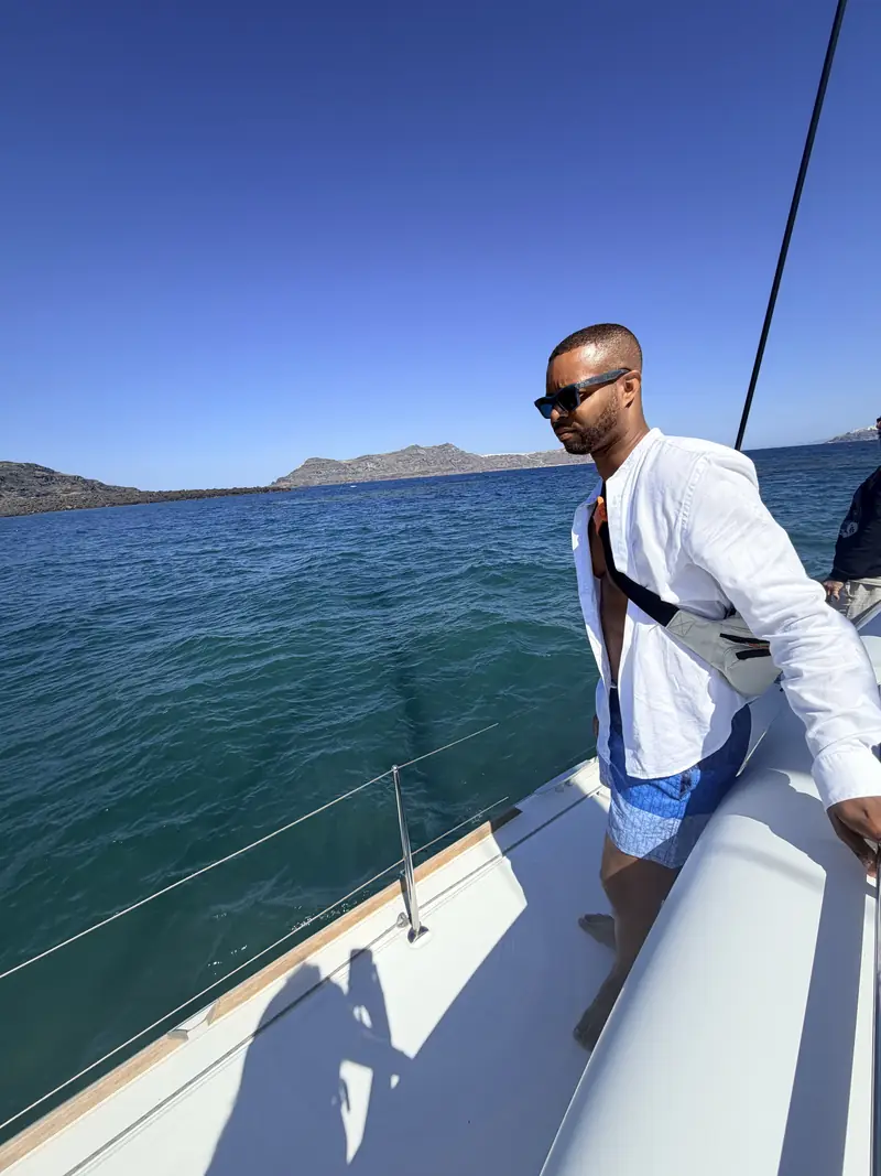Merrick Lee Jr. in a white open shirt and sunglasses standing on a sailboat deck, deep blue sea and distant islands behind him