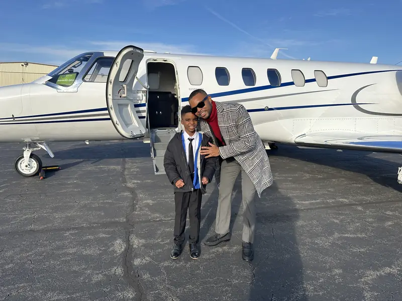 Merrick Lee Jr. and his son Maddox, both in suits, standing on the tarmac beside the open door of a private jet